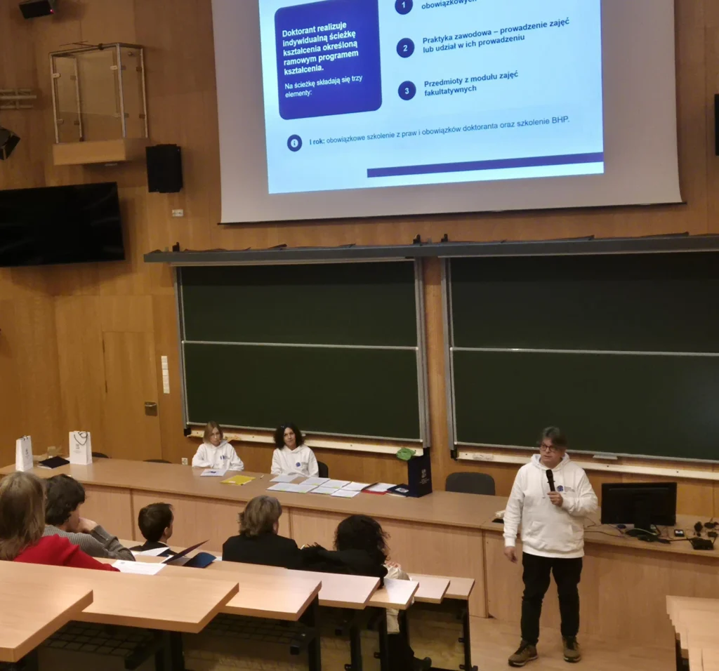 Prof. Radosław Fiedler z mikrofonem omawia ścieżkę kształcenia doktorantów; za nim slajd prezentacji i dwie osoby przy stole.